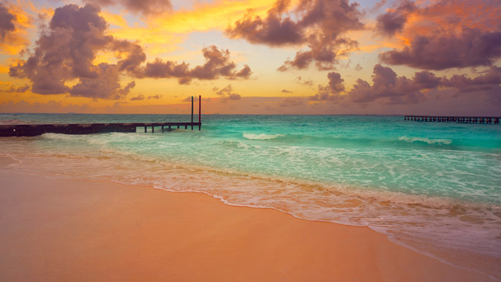 Playa Caracol, Cancún En primer plano se encuentra la playa de tono rosado, el agua del mar verde claro y la puesta de sol en colores rojizos